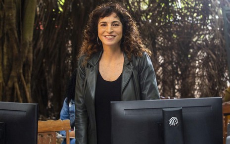De jaqueta e blusa pretas, Rosane Svartman posa em frente a duas telas de computador