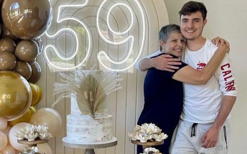 Rose Miriam abraçada ao filho João Augusto durante festa de aniversário; ambos estão ao lado de um neon luminoso com o número 59 e uma mesa com bolo branco e docinhos