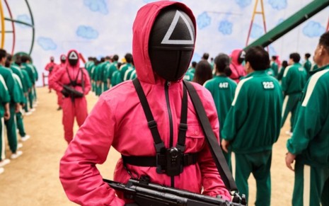 Pessoa fantasiada com macacão vermelho e máscara preto com um triângulo branco no meio e uma metralhadora na mão; cena da série Round 6