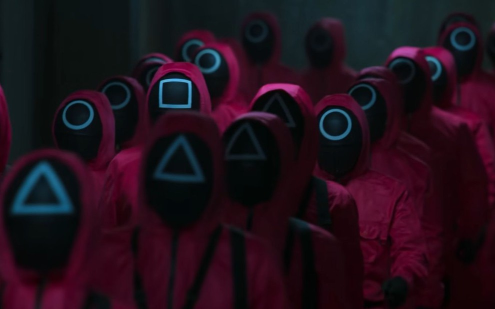 Imagem com cena da série Round 6, com pessoas vestidas com moletons vermelhos e roupas pretas com símbolos no rosto