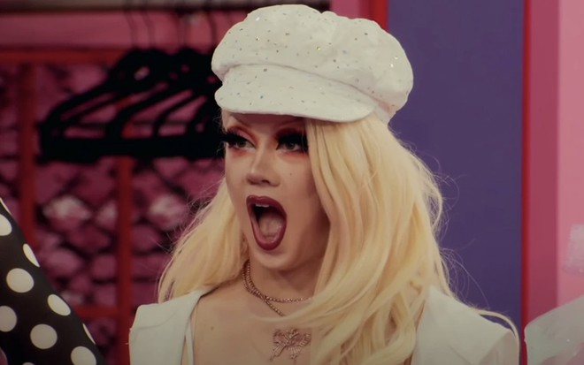 A drag queen Willow Pill de boca aberta, em reação de surpresa com uma peruca loira, uma boina branca, no Werk Room de Rupaul's Drag Race