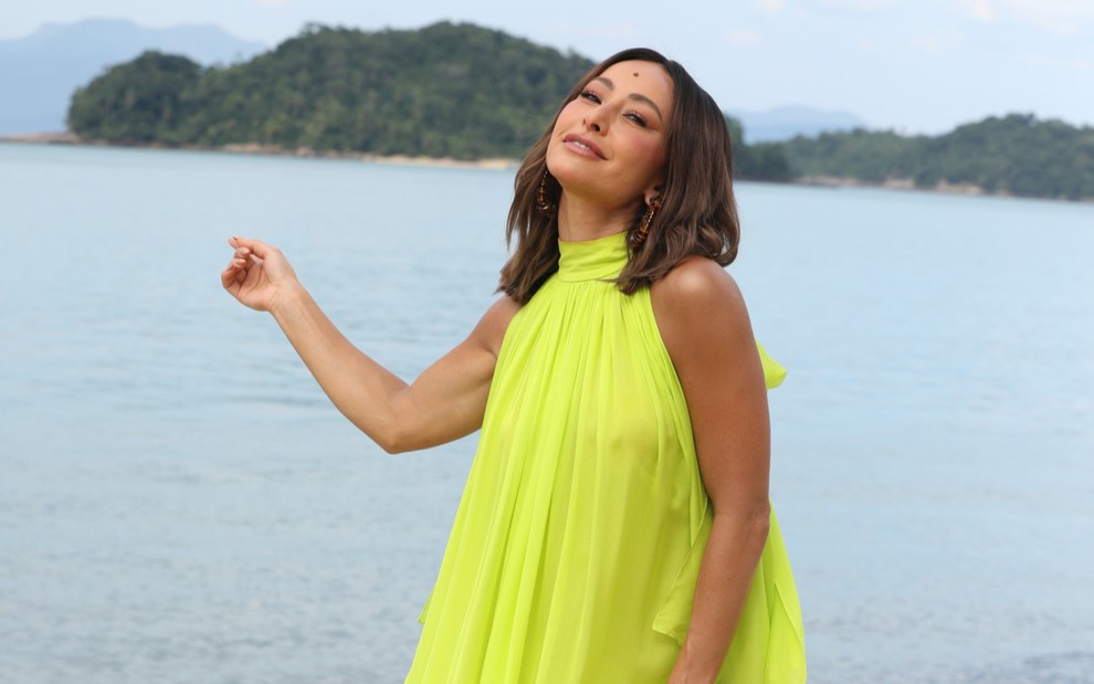 Sabrina Sato com um vestido verde limão, posando inclinada com o mar e uma ilha ao fundo