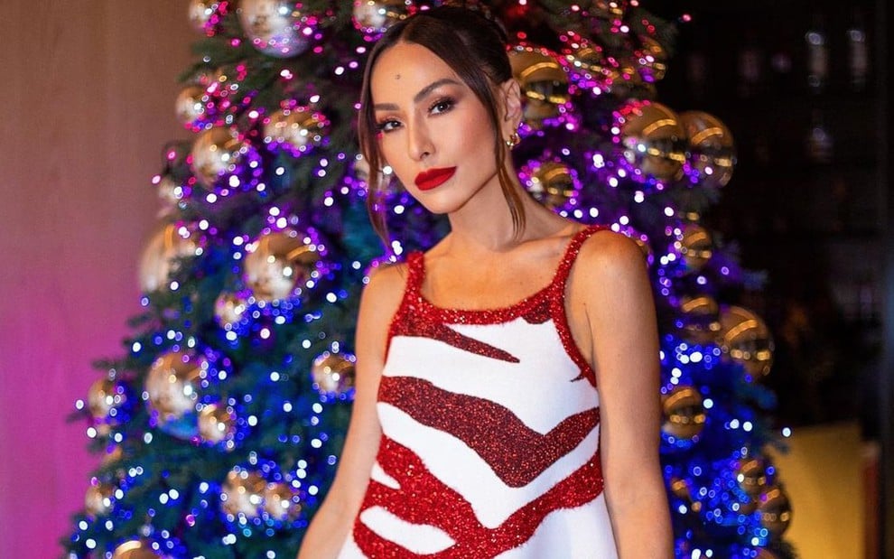 Com um vestido vermelho e branco, Sabrina Sato posa em frente a uma árvore de Natal