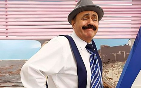 João Elias caracterizado como personagem Salim Muchiba. Ele usa suspensórios, gravata, bigode cheio e um chapéu