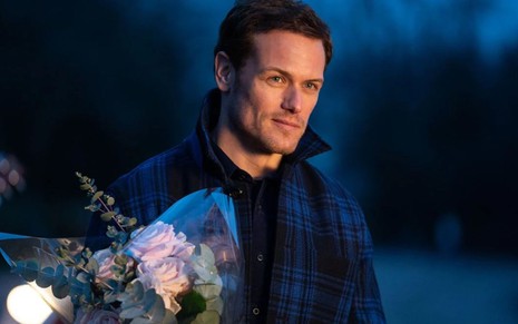 Sam Heughan caracterizado como Rob, seu personagem de O Amor Mandou Mensagem, com buquê de flores na mão
