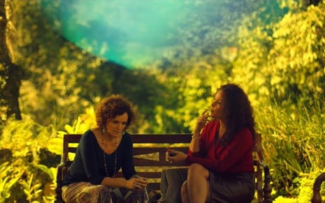 Sandra Corveloni e Carolina Ferraz sentadas em um banco a céu aberto; no fundo há um cenário verde e uma forte luz. Sandra está com os cotovelos apoiados nos joelhos e Carolina está fumando.