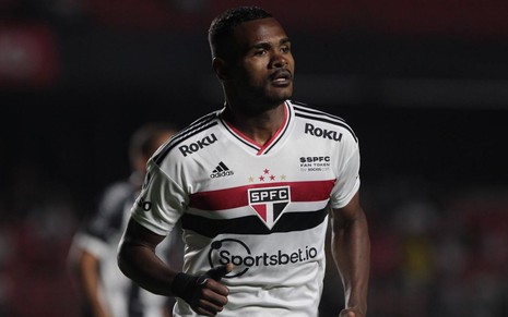 Jogador Nikão, do São Paulo, corre em campo e veste uniforme branco com detalhes pretos e vermelhos