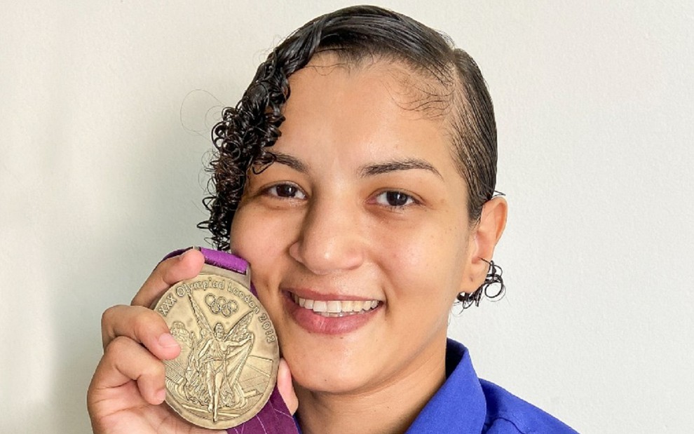 Sarah Menezes com uma blusa azul e sua medalha de ouro nos Jogos Olímpicos de Tóquio na mão, ao sorrir para a câmera