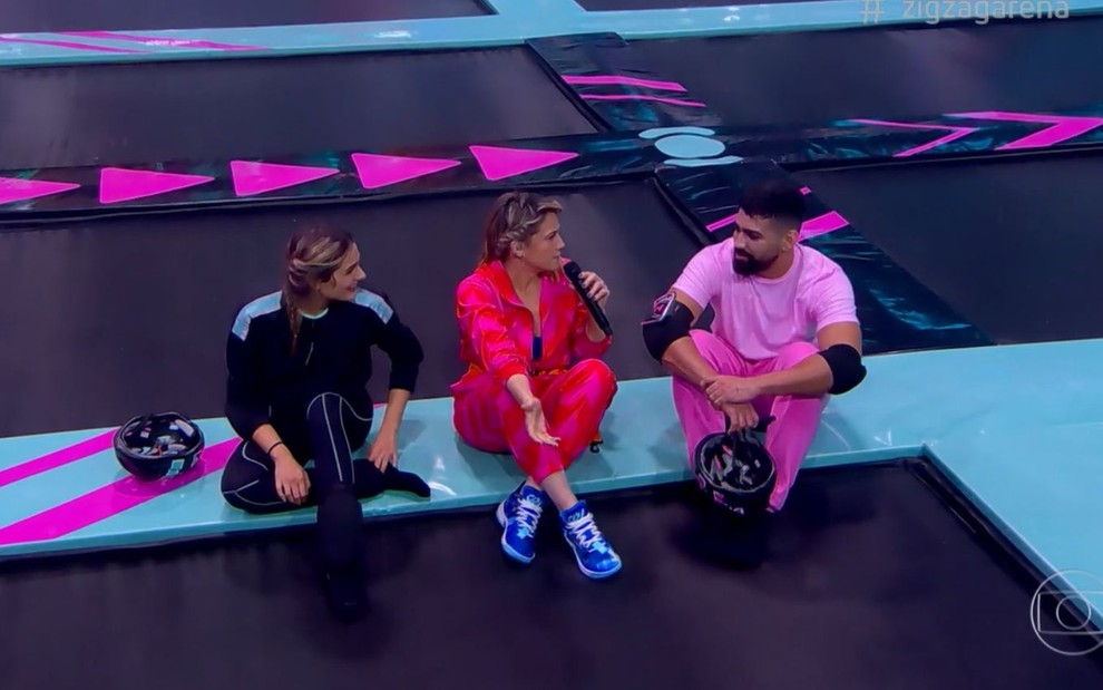 Sasha, Fernanda Gentil e Dilsinho sentados sobre uma estrutura verde centralizada em uma cama elástica gigante