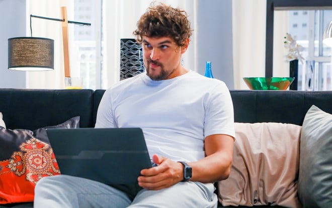 Mauro (Felipe Roque) com um notebook no colo sentado no sofá em cena de A Infância de Romeu e Julieta