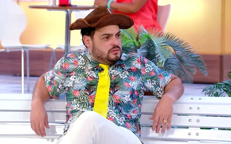 Matheus Ceará com chapéu de cangaceiro, camisa florida e gravata amarela sentado no banco da praça participação em A Praça É Nossa, do SBT