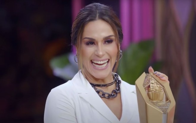 A apresentadora Nadja Haddad no Bake Off Brasil, programa exibido pelo SBT; ela sorri, animada, segurando um troféu do reality