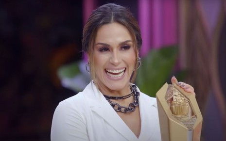 A apresentadora Nadja Haddad no Bake Off Brasil, programa exibido pelo SBT; ela sorri, animada, segurando um troféu do reality
