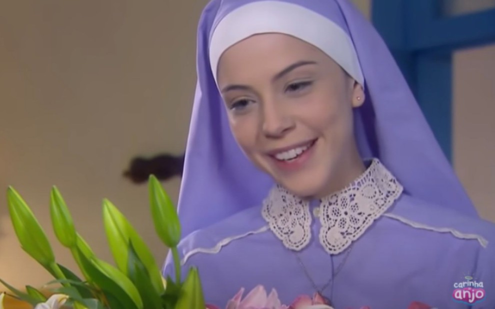 Bia Arantes grava cena sorrindo segurando flores, como Irmã Cecília em Carinha de Anjo