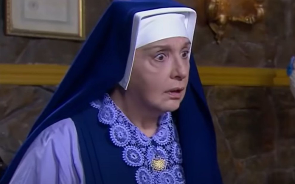 Eliana Guttman grava cena vestida de freira, com expressão séria, como Madre Superiora em Carinha de Anjo, do SBT