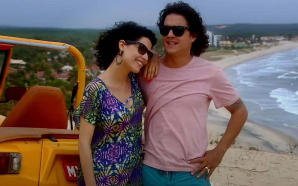 Manuela do Monte e Guilherme Boury gravam cena abraçados e sorrindo, com a praia de fundo da imagem, como Carol e Junior