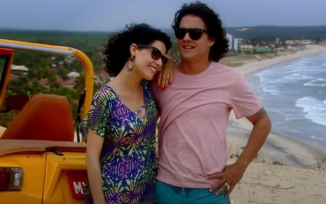 Manuela do Monte e Guilherme Boury gravam cena abraçados e sorrindo, com a praia de fundo da imagem, como Carol e Junior