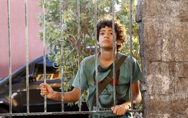 Gabriel Santana grava apoiado em grade com camiseta verde e cara desanimada como Mosca de Chiquititas