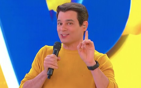 O apresentador Celso Portiolli aponta com o dedo indicador da mão direita para cima e segura um microfone com a mão esquerda no palco do Domingo Legal