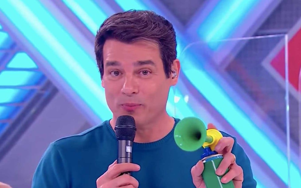 O apresentador Celso Portiolli segura um microfone com a mão esquerda e uma buzina verde e amarela com a mão direita no palco do Domingo Legal