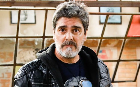Junno Andrade, caracterizado como Renato, em ensaio de divulgação de Poliana Moça; ele tem o cabelo e a barba grisalhas e ergue as sobrancelhas enquanto encara a câmera.