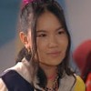 Bella Chiang, caracterizada como Song, dá um leve sorriso; ela tem o cabelo preso em um coque e veste o uniforme da escola Ruth Goulart em cena de Poliana Moça
