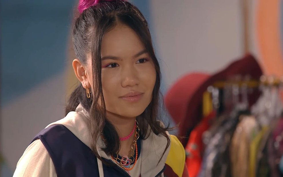 Bella Chiang, caracterizada como Song, dá um leve sorriso; ela tem o cabelo preso em um coque e veste o uniforme da escola Ruth Goulart em cena de Poliana Moça
