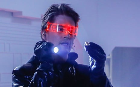 Pedro Lemos, caracterizado como Waldisney, usa óculos transparente com LED vermelho e roupa preta. As mãos, com luvas, seguram uma lanterna acesa em cena de Poliana Moça.