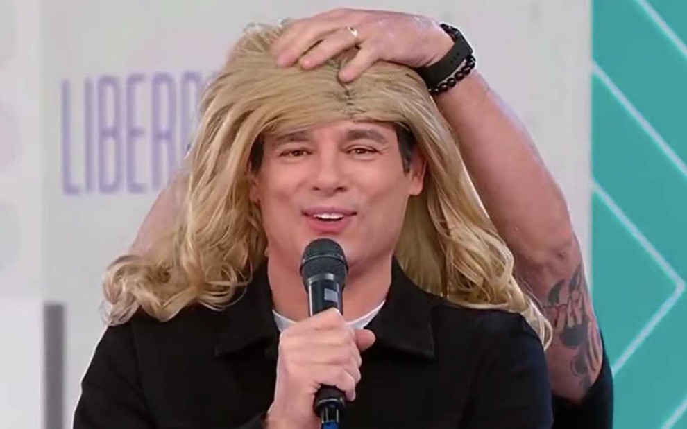 O apresentador Celso Portiolli faz cara de vergonha enquanto uma pessoa coloca uma peruca loira na sua cabeça no Teleton