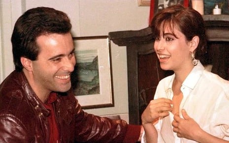 Os atores Tony Ramos e Fernanda Torres como Cristiano e Simone em Selva de Pedra; eles estão frente a frente, enquanto ele ri e coloca a mão nela, que também ri com as mãos no peito, mexendo na roupa