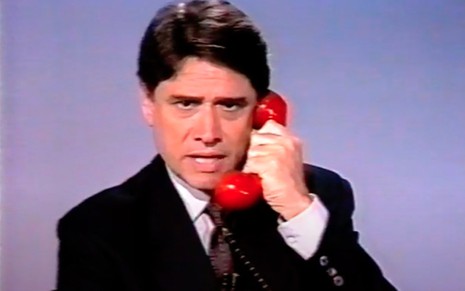 O jornalista Sérgio Chapelin atendendo a um telefone com fio vermelho, de terno preto, no estúdio do Jornal Nacional em 1991