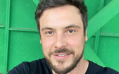 Sergio Guizé sorri e está em frente a uma parede verde