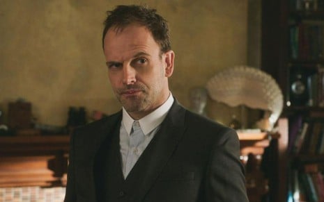 Sherlock (Jonny Lee Miller) de camisa branca e paletó preto olhando para a direita em sala com detalhes de decoração no fundo em cena de Elementary