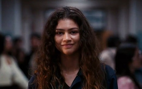 Rue (Zendaya) de moletom em corredor de um colégio dando um sorriso em cena do teaser da segunda temporada de Euphoria