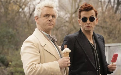 Aziraphale (Michael Sheen) no lado esquedo de roupa branca com sorvete na mão e Crowley (David Tennant) de roupa preta e óculos escuros com picolé na mão em cena da série Good Omens