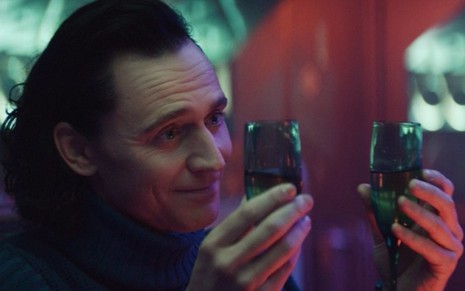 Loki (Tom Hiddleston) de blusa azul e com duas taças com bebida seguradas em cada mão olhando para a direita em cena da série Loki