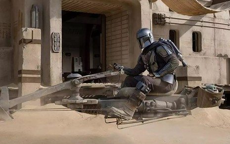 O Mandalorian de armadura prateada em cima de moto espacial voadora e com um alienígena verde ao seu lado em cena de The Mandalorian, do Disney+