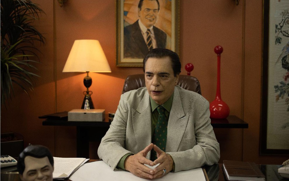 Silvio Santos (José Rubens Chachá) de camisa verde e paletó claro sentado em mesa em um escritório com as mãos se tocando em cena da série O Rei da TV