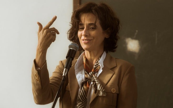 Natália (Mariana Lima) com camisa branca, jaqueta marrom e lenço no pescoço fala em frente a um microfone com a mão direita levantada em cena da série Os Dias Eram Assim (2017)