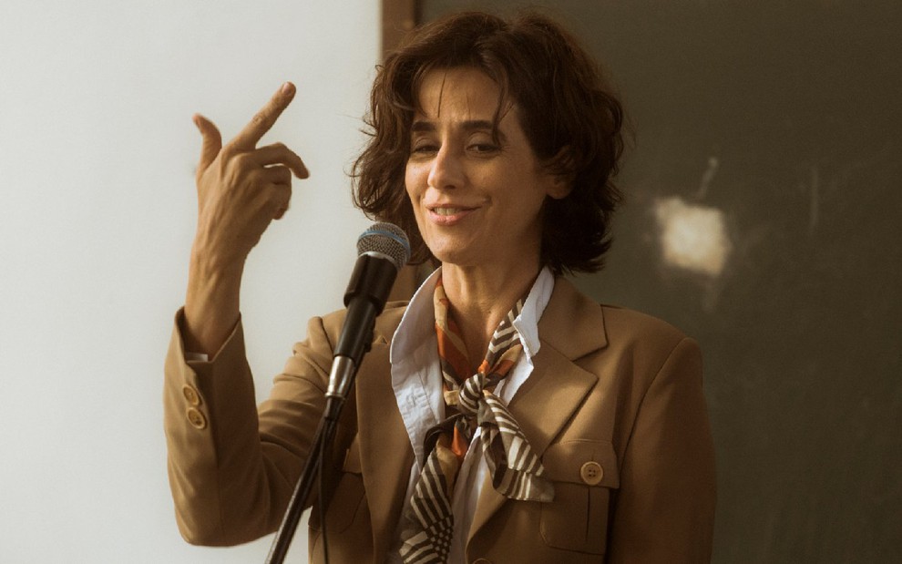 Natália (Mariana Lima) com camisa branca, jaqueta marrom e lenço no pescoço fala em frente a um microfone com a mão direita levantada em cena da série Os Dias Eram Assim (2017)