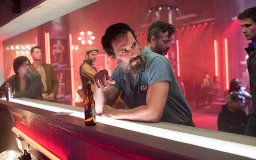 Joel (Rodrigo Santoro) encostado em balcão de bar com uma cerveja na frente e o braço direito levantado olhando para a direita com personagens ao fundo em cena da série Reprisal