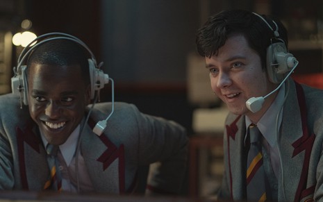 Eric (Ncuti Gatwa) no lado esquerdo e Otis (Asa Butterfield) no lado direito sorrindo com uniformes cinzas e fones de ouvido branco em cena da terceira temporada de Sex Education