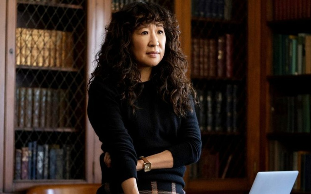 Dr. Ji-Yoon Kim (Sandra Oh) de blusa preta e braços cruzados diante de fundo com livros espalhados por prateleiras em cenas da série The Chair