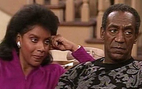 Phylicia Rashad no lado esquerdo de roupa roxa e Bill Cosby no lado direito de blusa escura com detalhes claros sentados em um sofá olhando para a direita em cena da série The Cosby Show
