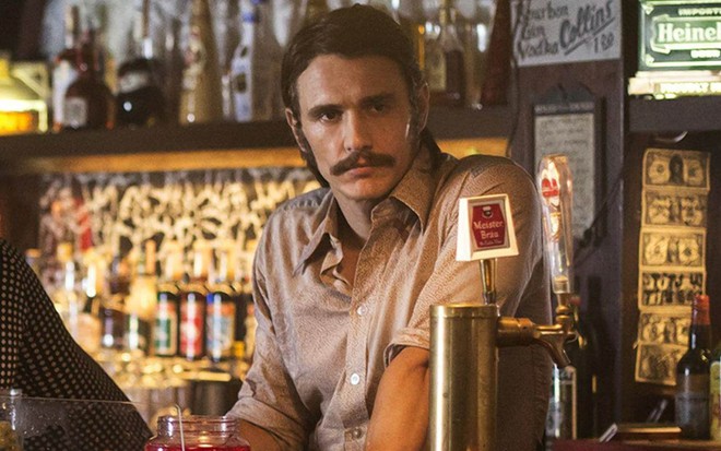 James Franco no papel de Vincent Martino com camisa cinza e bigode atrás de um balcão de bar em cena da série The Deuce