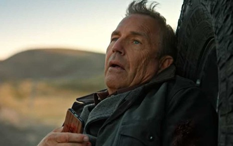 John Dutton (Kevin Costner) encostado em roda do carro de jaqueta escura olhando para o alto com o celular na mão direita e marca de tiro no braço esquerdo em cena da série Yellowstone