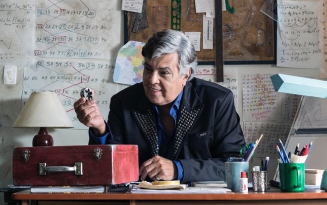 Sidney Magal sentado em uma escrivaninha, com um braço apoiado na mesa e a outra mão levantada segurando um pequeno objeto