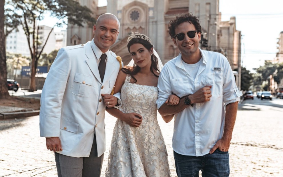 Sidney veste um terno branco e uma gravata escura, ele está com o braço enganchado em Antônia Morais, que está ao centro vestida de noiva; à direita está Rafael Almeida, com uma camisa branca, óculos escuros e sorrindo