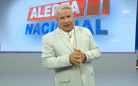 Sikêra Jr. com as mãos cruzadas, vestindo um terno cinza claro no cenário de seu telejornal