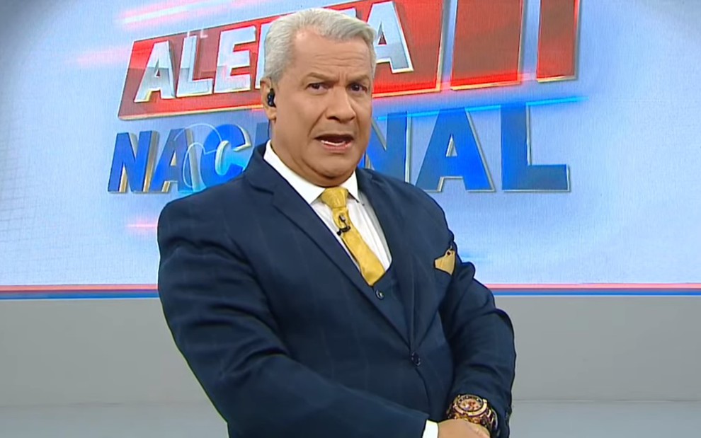 Sikêra Jr. de braços cruzados, terno azul, gravata amarela e camisa branca no cenário do Alerta Nacional
